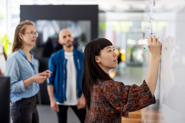  Team of three coworkers in a brainstorm meeting with one person writing on a whiteboard.