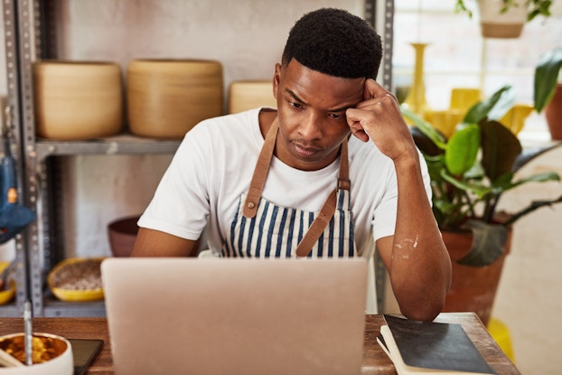  A man sits at a table in front of an open laptop with a worried, tired expression. He wears a striped apron over a white T-shirt and has his head leaning on one hand. Behind him is a metal shelving unit filled with large pots. A potted plant sits on the floor next to the shelves.