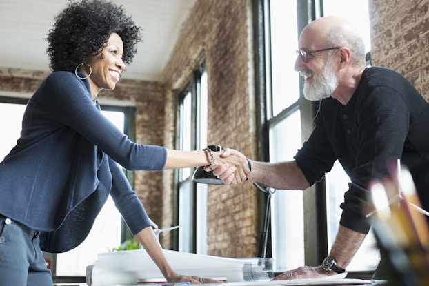 A woman wearing a black blazer and large hoop earrings and a white-bearded man wearing a black long-sleeved shirt shake hands across a table.