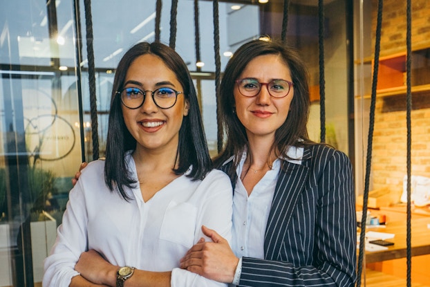 Two young female business partners stand next to each other while smiling at the viewer. Behind them is a modern office.