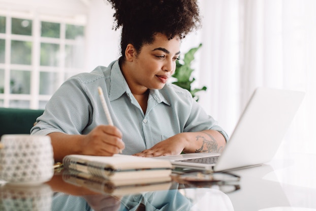 A woman is sitting at her kitchen table in a sunlit room in front of a laptop computer. She is looking at her computer and writing notes in a notebook.