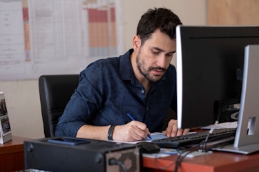 A man is seated at a desk in an office. He sits before a desktop computer. He has a pen in his right hand and he is making notes on a sheet of paper. He is doing a careful analysis of his business plan.