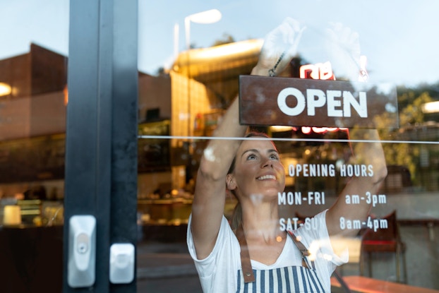  Smiling woman holding up an "open" sign on the door to her business.