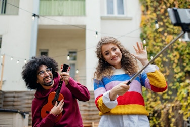  A woman in a multicolored striped sweater uses a smartphone on a selfie stick to film herself and a young man who plays a ukelele. 