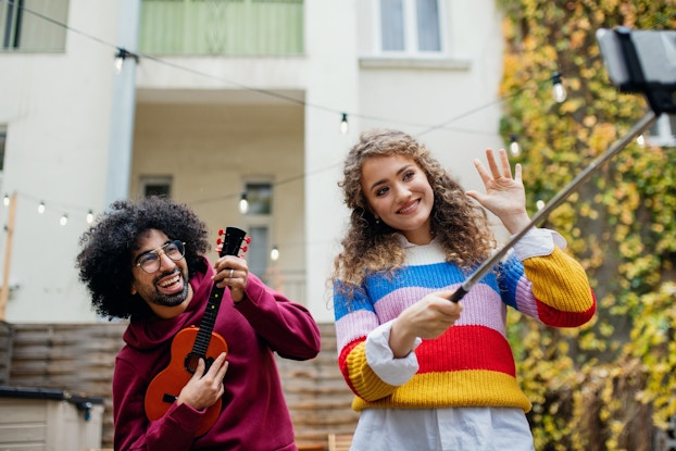  A woman in a multicolored striped sweater uses a smartphone on a selfie stick to film herself and a young man who plays a ukelele.