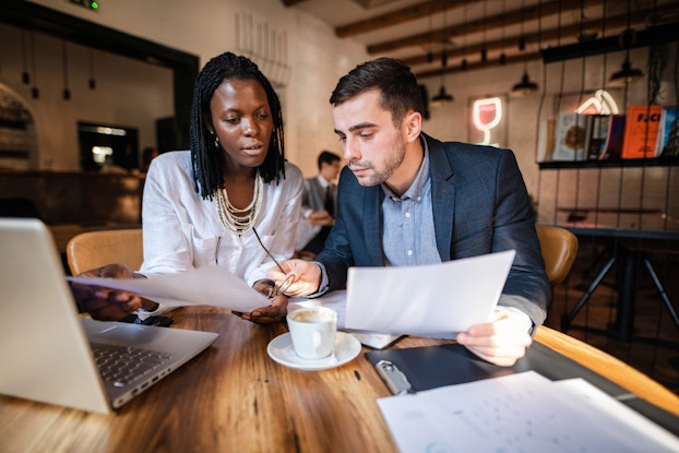A woman restaurant owner is seated at a table in her cafe with a business valuator. They are reviewing spreadsheets and financial data to determine the worth of her establishment.