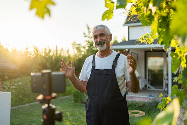  A man stands outside, in front of a farmhouse and films himself speaking. In the foreground, slightly out of focus, a smartphone has been mounted horizontally on a tripod. The man is older, with salt-and-pepper hair and a white beard. He wears overalls over a white T-shirt. He is smiling broadly and standing with his hands up and slightly spread. The house behind him is white with a gray roof and a storm door with a full glass screen. Next to the farmhouse, the sun rises over tall rows of crops.