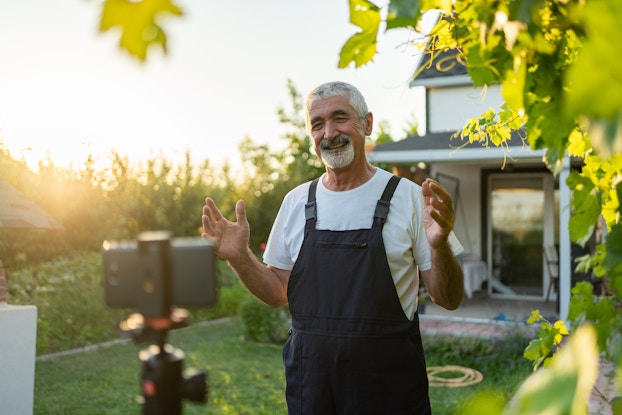 A man stands outside, in front of a farmhouse and films himself speaking. In the foreground, slightly out of focus, a smartphone has been mounted horizontally on a tripod. The man is older, with salt-and-pepper hair and a white beard. He wears overalls over a white T-shirt. He is smiling broadly and standing with his hands up and slightly spread. The house behind him is white with a gray roof and a storm door with a full glass screen. Next to the farmhouse, the sun rises over tall rows of crops.