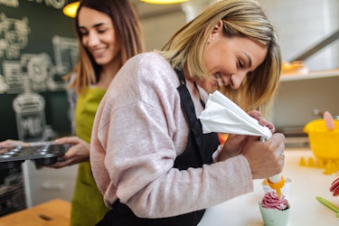  Two women work in the kitchen of a cupcake shop. The first woman, who has shoulder-length blonde hair, uses an icing bag to swirl pink icing on top of a cupcake. The second woman, who has brown hair, walks behind her coworker with an empty cupcake tin. 