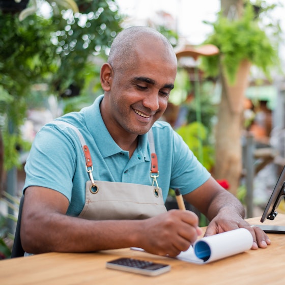 A bald man wearing a teal collared shirt under a tan apron sits at a wooden table outside. He smiles as he writes something on a notepad. Beside the notepad on the table are a smartphone on a stand and a small calculator. Behind the man are lines of potted plants and a cluster of trees, with an out-of-focus building standing a greater distance away.