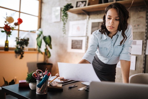 A business woman leans over her desk. She is holding up a paper in her left hand. She is reviewing an application as she prepares to finalize incorporation paperwork with her state government.