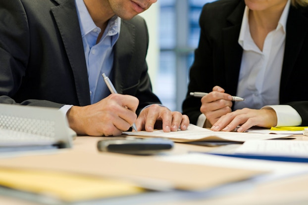 A close-up photo shows a man (his face is not visible) signing a contract. There is a woman seated next to him that is holding a pen and is prepared to sign the contract as well.
