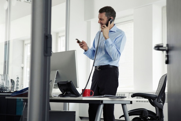 A businessman is using his company's landline to speak with a client on the phone. He is holding a cellphone in the other hand is checking messages while talking on the phone.