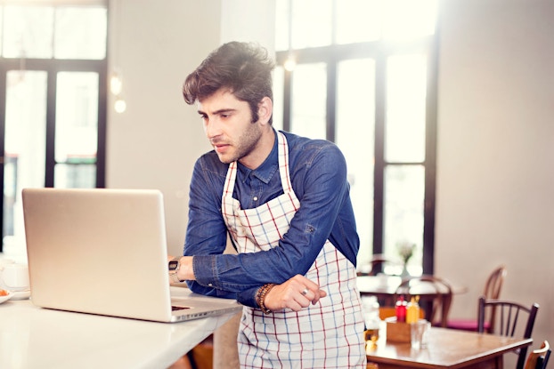  A young cafe owner stands before a laptop computer resting on a counter in his restaurant. The man is looking at his laptop with a concerned look on his face.