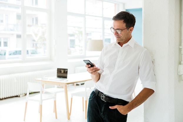A businessman looks on his cell phone. Behind him is a table and laptop computer sitting in a bright office.