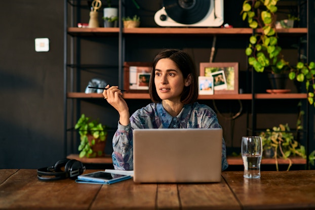 A woman entrepreneur working from home holds a pen and contemplates something. She is seated at a table with a laptop before her.