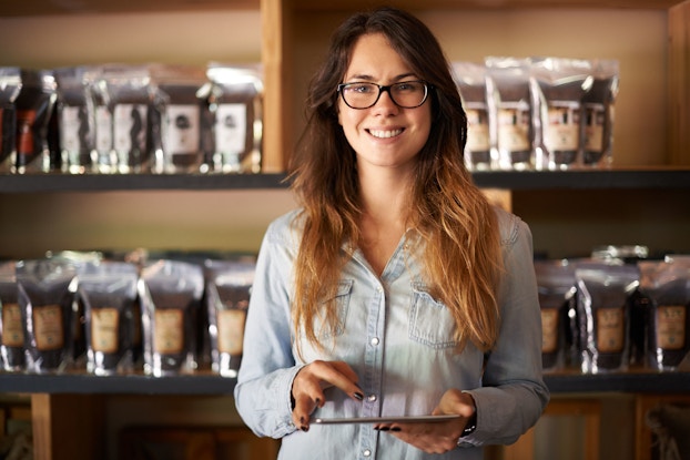  A woman business owner of a coffee roasting establishment smiles at the viewer. She is holding a computer tablet in her hand.