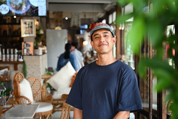  A cafe owner proudly stands in his restaurant facing the camera smiling.