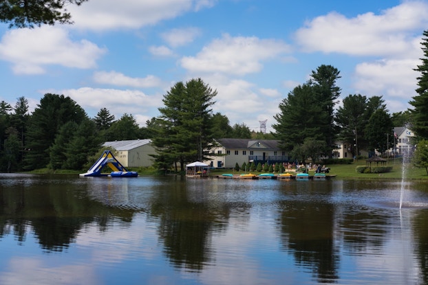  The lake at Camp Kennybrook in Monticello, New York. Across the lake can be see several buildings among pine trees, a blue-and-yellow inflatable water slide and several blue and yellow rowboats next to a wooden dock.