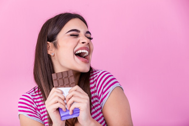  Woman excitedly smiling while holding a chocolate bar in front of a hot pink background.