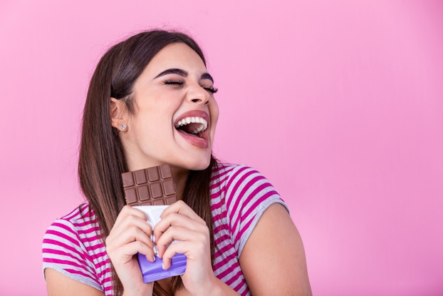 Woman excitedly smiling while holding a chocolate bar in front of a hot pink background.