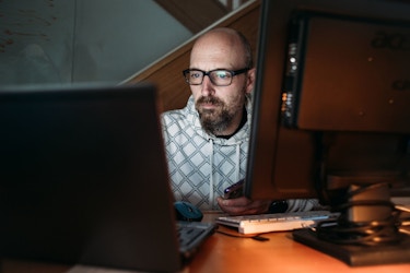  A man with glasses works at a desktop computer and a laptop late at night. He is looking at his laptop screen intently. 
