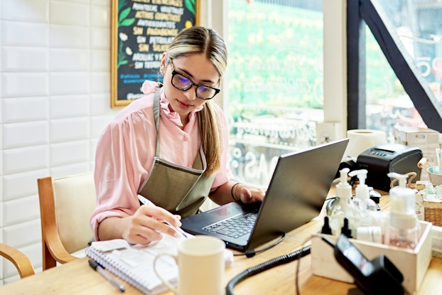A young woman sits at a desk with an open laptop and a notebook in front of her. She uses her right hand to write something in the notebook and has her left hand on the keyboard of the laptop. The woman has long blonde hair pulled into a ponytail and she wears glasses and a pale pink blouse under an olive green apron. Also on the desk are baskets of hand soaps and lotions and a receipt printer.