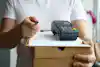 A delivery man pictured from the chest below is holding multiple boxes with a clipboard and invoice and mobile point-of-sale unit resting on top of the boxes. He is holding a pen for the customer to sign the invoice.