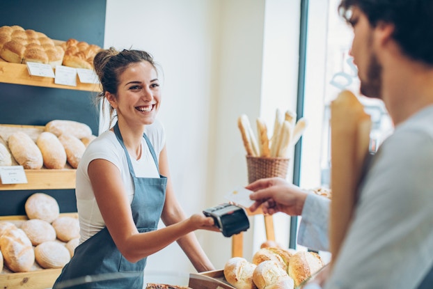  A woman stands behind the counter at a bakery and holds out a handheld credit card scanner so the customer across the counter can scan his card. The woman has dark blonde hair pulled into a bun and she wears a navy blue apron over a white T-shirt. She is smiling broadly at the customer, who is out of focus in the foreground and partially offscreen. The checkout counter is covered with loaves of bread. Bread also fills angled shelves on the wall behind the counter.