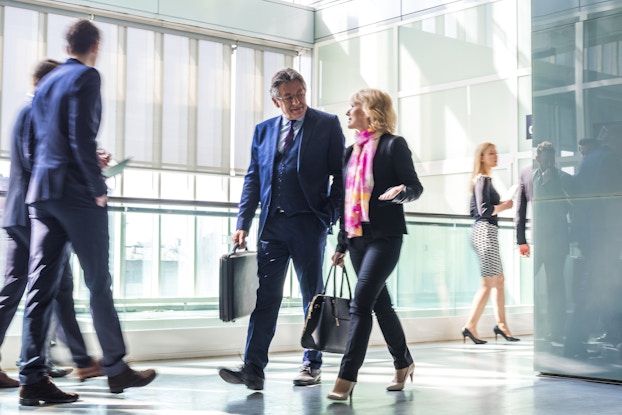  A pair of businesspeople walks through a hallway in a brightly lit modern building. Several other businesspeople are in motion around them. In the background are floor-to-ceiling, wall-to-wall glass windows.