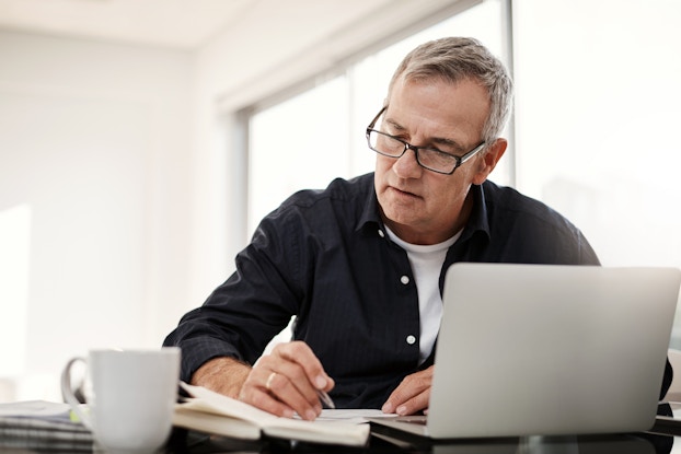 An older man wearing glasses sits at a table in front of an open laptop. He's looking down at and writing on a paper. Next to the paper and the laptop on the table are an open book, a coffee mug, and a stack of newspapers.