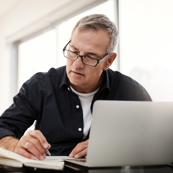 An older man wearing glasses sits at a table in front of an open laptop. He's looking down at and writing on a paper. Next to the paper and the laptop on the table are an open book, a coffee mug, and a stack of newspapers.