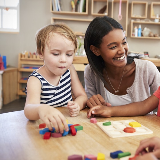 A woman sits between two children of about 3 or 4 years of age around a low wooden table. The child on the left is grabbing at one of the 3D shapes in the pile in front of him. The child on the right is grinning at the adult woman, who smiles widely back at him. This child has a puzzle in front of him with nine spaces for nine different shapes. Three of the spaces are filled with shapes (octagon, semicircle, and square) from the pile on the table. In the foreground, a third child looks down at his own pile of shapes.