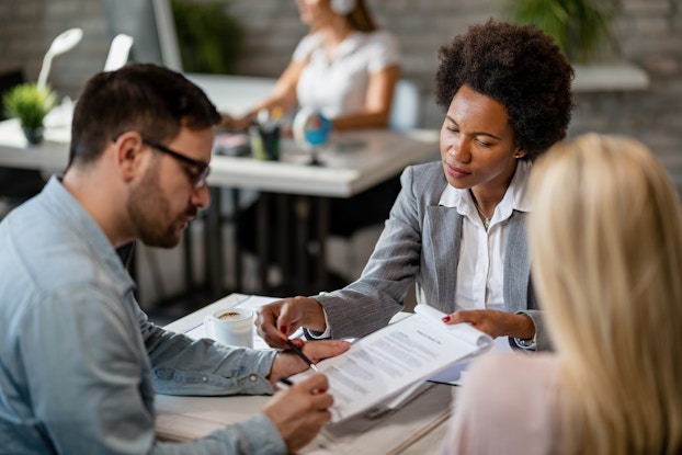 A woman in a gray blazer and white collared shirt sits across a desk from a couple and holds out a contract. The man sitting on the left signs the contract. The woman on the right looks on, facing away from the camera. In the background, out of focus, another woman sits and works at another desk.