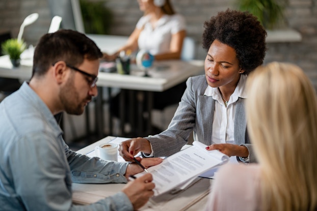  A woman in a gray blazer and white collared shirt sits across a desk from a couple and holds out a contract. The man sitting on the left signs the contract. The woman on the right looks on, facing away from the camera. In the background, out of focus, another woman sits and works at another desk.