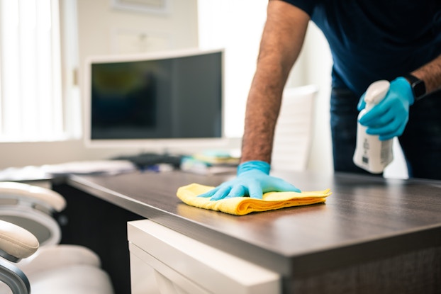 A person wearing light blue latex gloves uses a spray bottle and a yellow cloth to clean the top of a desk. The person's head and shoulders are out of frame. A room with large windows and a wide-screen TV can be seen out-of-focus in the background.