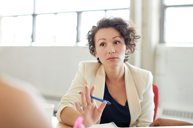 A woman with short curly hair sits at a table and faces the person across from her with a serious expression and her eyebrows raised. She has one arm on the table, the hand raised as if asking for patience. The shoulder of the person across from the woman can be seen in the foreground, out of focus.