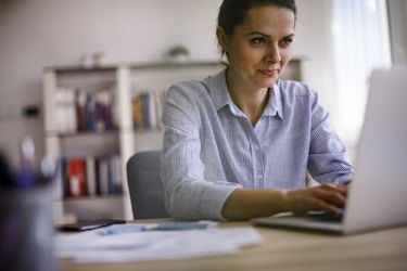 A young woman in a button-up shirt works on a laptop. Behind her, out of focus, is a book-lined set of shelves, and there are papers and pens on the desk beside her.