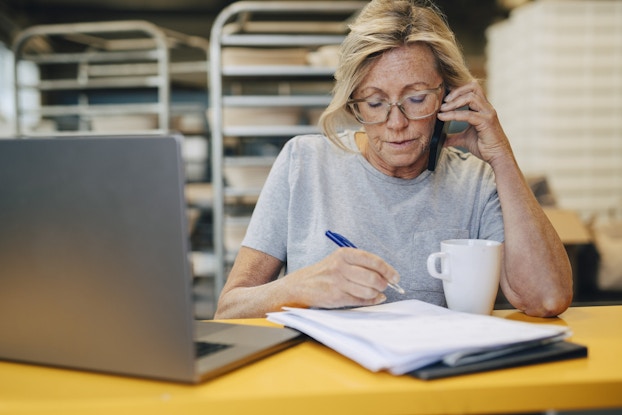A middle-aged woman sits at a counter in the back room of a bakery. She's talking on a smartphone while writing something in a notebook. The woman has blonde hair pulled back and she's wearing a light gray T-shirt and glasses. An open laptop sits in front of her on the counter, beside her notebook. Behind her are steel shelves holding trays and dishes.