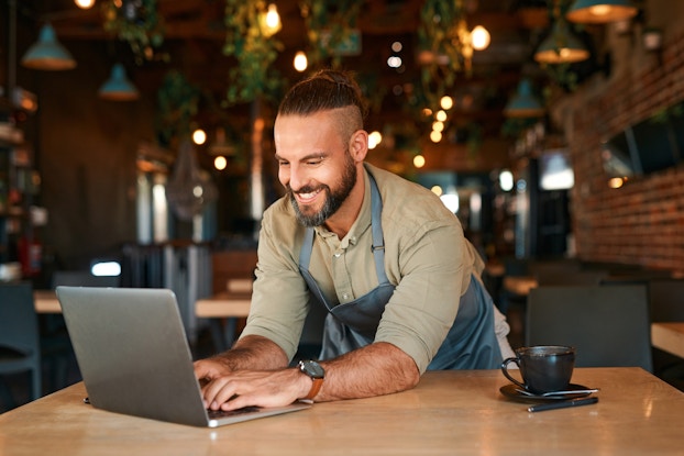A smiling man leans over a wood-topped table and types on the keyboard of the laptop open in front of him. The man wears a blue apron over a khaki button-up shirt, and he has dark hair and a beard. In the background, out of focus, is an empty restaurant filled with more wood-topped tables and black chairs. Plants are entwined with the lighting fixtures that hang from the ceiling.
