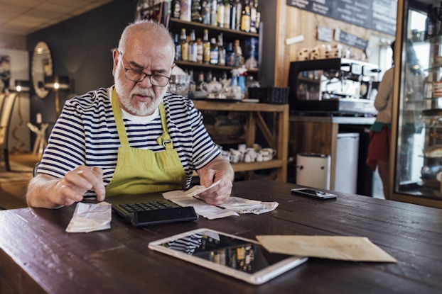  An older man sits at a rustic wooden table and reviews receipts. His brow is furrowed as he uses a finger to locate something on one of the receipts. The receipts look slighty crumpled and are arranged in three rows on the table, with a large black calculator between two of the rows. The man has a white beard and wears glasses and a neon green apron over a black-and-white striped shirt. He appears to be sitting in a cafe or restaurant; in the background are shelves of bottles and mugs and a large coffee machine.