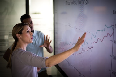  A woman and a man stand in front of a large screen displaying a chart labveled "Price Index/Earnings." Two jagged lines, one blue and one red, rise from the lower left side of the chart to the upper right, showing growth. The woman stands closer to the viewer and uses her right hand to gesture to the right side of the chart. The man stands with his left hand up and open. 