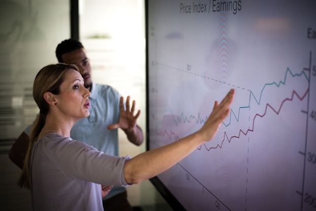 A woman and a man stand in front of a large screen displaying a chart labveled "Price Index/Earnings." Two jagged lines, one blue and one red, rise from the lower left side of the chart to the upper right, showing growth. The woman stands closer to the viewer and uses her right hand to gesture to the right side of the chart. The man stands with his left hand up and open.