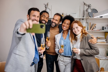  A group of five people in business casual clothes stand close together in a line and smile for a selfie. The camera phone is held horizontally by the man on the far left of the group. Everyone in the group holds a glass of champagne, except for the man in the very middle, whose arms are stretched behind the people on either side of him. 