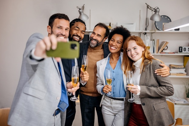 A group of five people in business casual clothes stand close together in a line and smile for a selfie. The camera phone is held horizontally by the man on the far left of the group. Everyone in the group holds a glass of champagne, except for the man in the very middle, whose arms are stretched behind the people on either side of him.