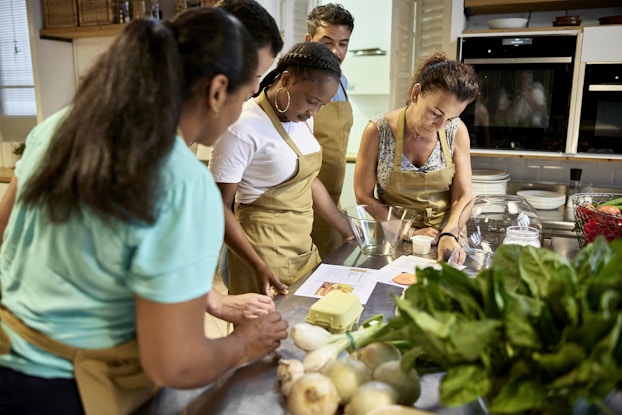 Five people wearing khaki aprons stand around the left side of a metal-topped table in a kitchen. They're looking down at a couple of recipe printouts. Also on the table are a couple of glass bowls, some white and green onions, a large amount of leafy greens, and a bowl of various other vegetables.