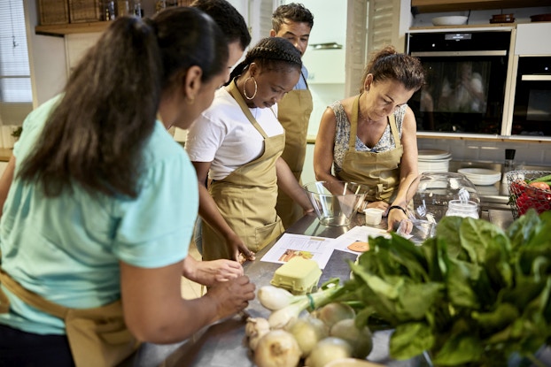  Five people wearing khaki aprons stand around the left side of a metal-topped table in a kitchen. They're looking down at a couple of recipe printouts. Also on the table are a couple of glass bowls, some white and green onions, a large amount of leafy greens, and a bowl of various other vegetables.