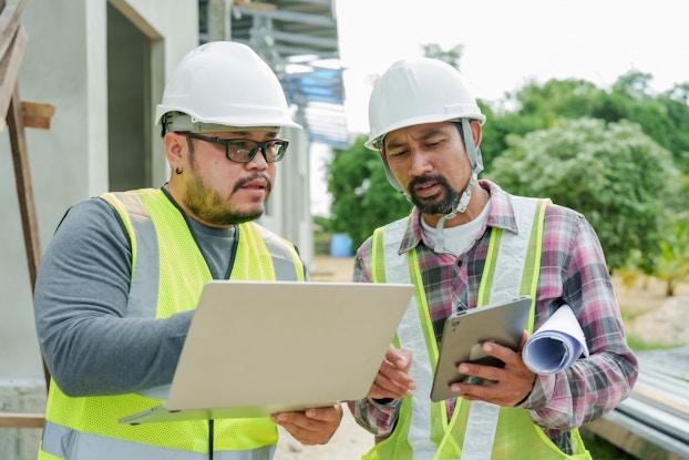 Two men in high-visibility vests and white hard hats stand outside next to a building. The man on the left wears glasses and holds an open laptop. The man on the right has a goatee and holds an electronic tablet. He points at something on the laptop's screen.