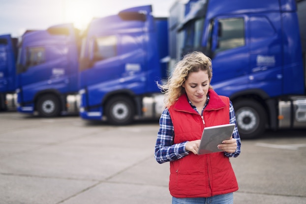  A woman stands outside, using an electronic tablet. Behind her is a row of parked 18-wheel trucks with blue cabins. The woman has blonde hair blowing in the wind and she wears a red puffed vest over a blue plaid shirt.