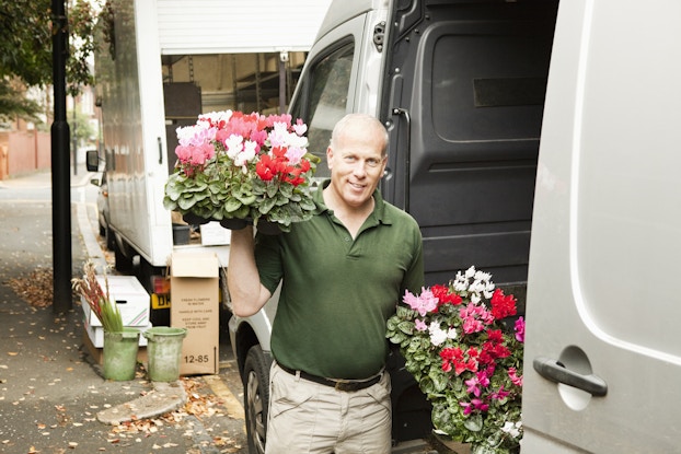  A man stands next to the open sliding door of a large white van, holding two bouquets of flowers. The man is bald and wearing a dark green polo shirt and khakis. The bouquets are made up of clusters of white, pink, and red flowers.